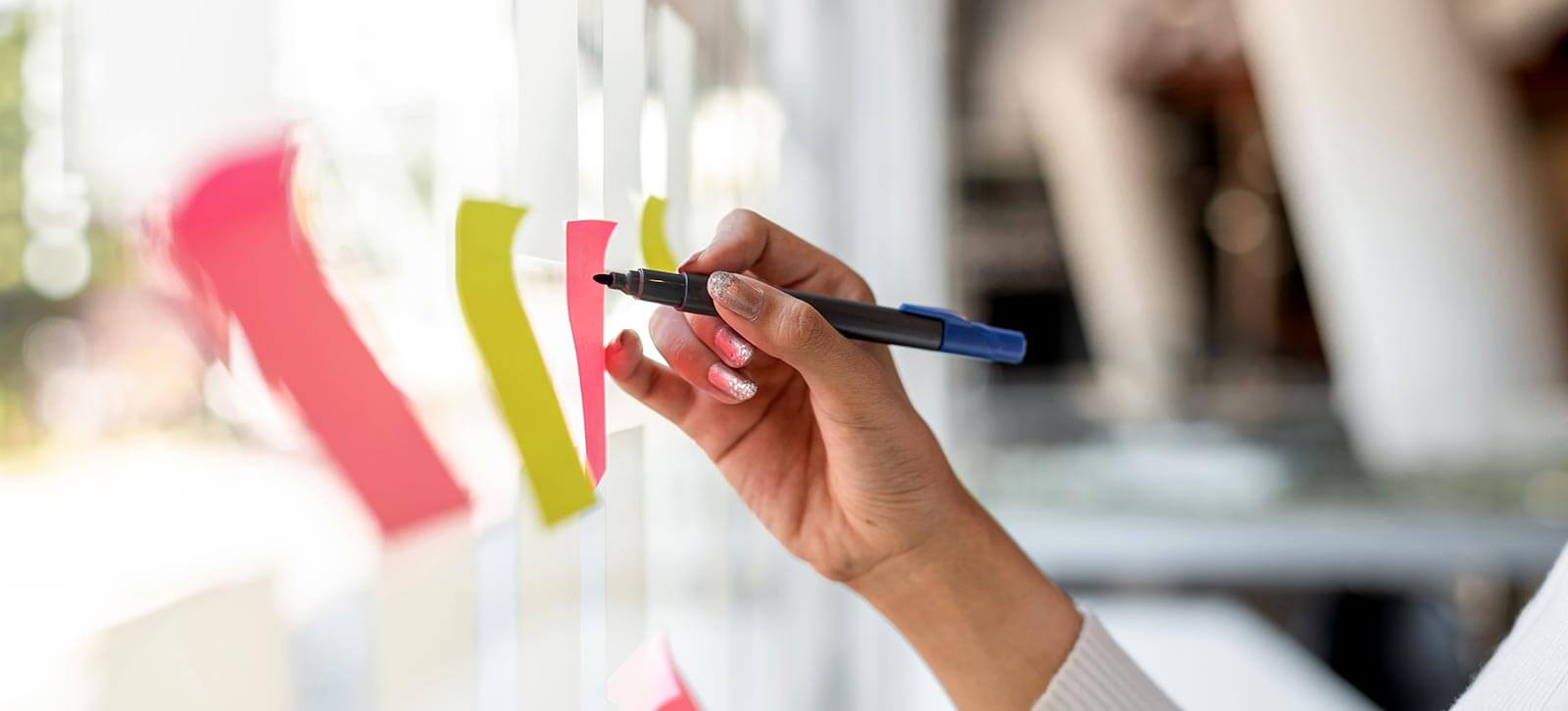 Close up of a person's hand writing on a glass wall filled with post it notes