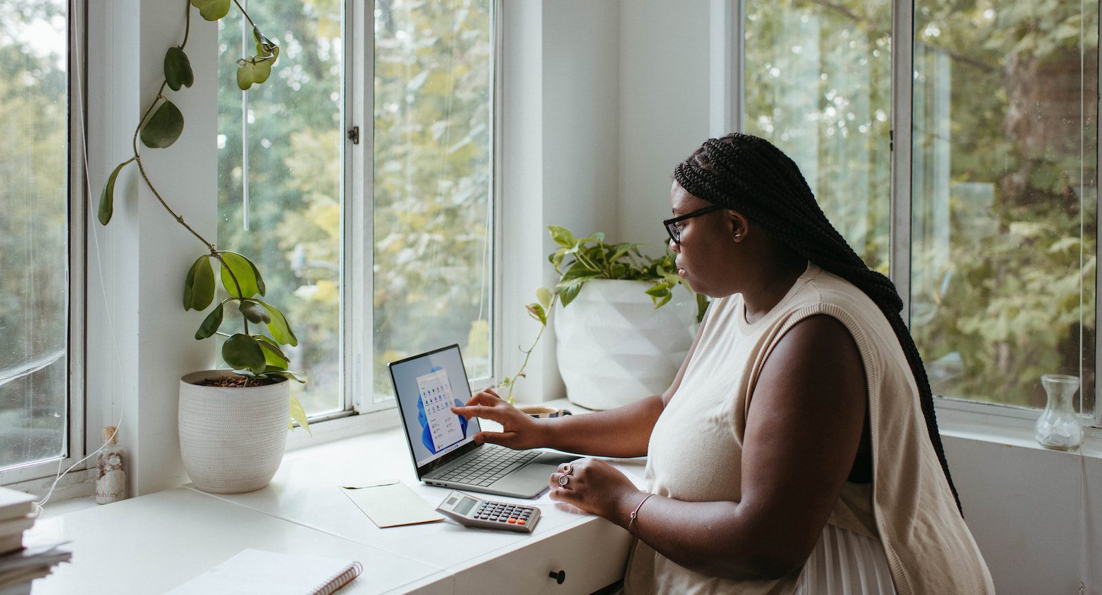 a woman sitting at a table in a home office with a laptop open in front of large windows revealing a bright, tree-filled forest outside